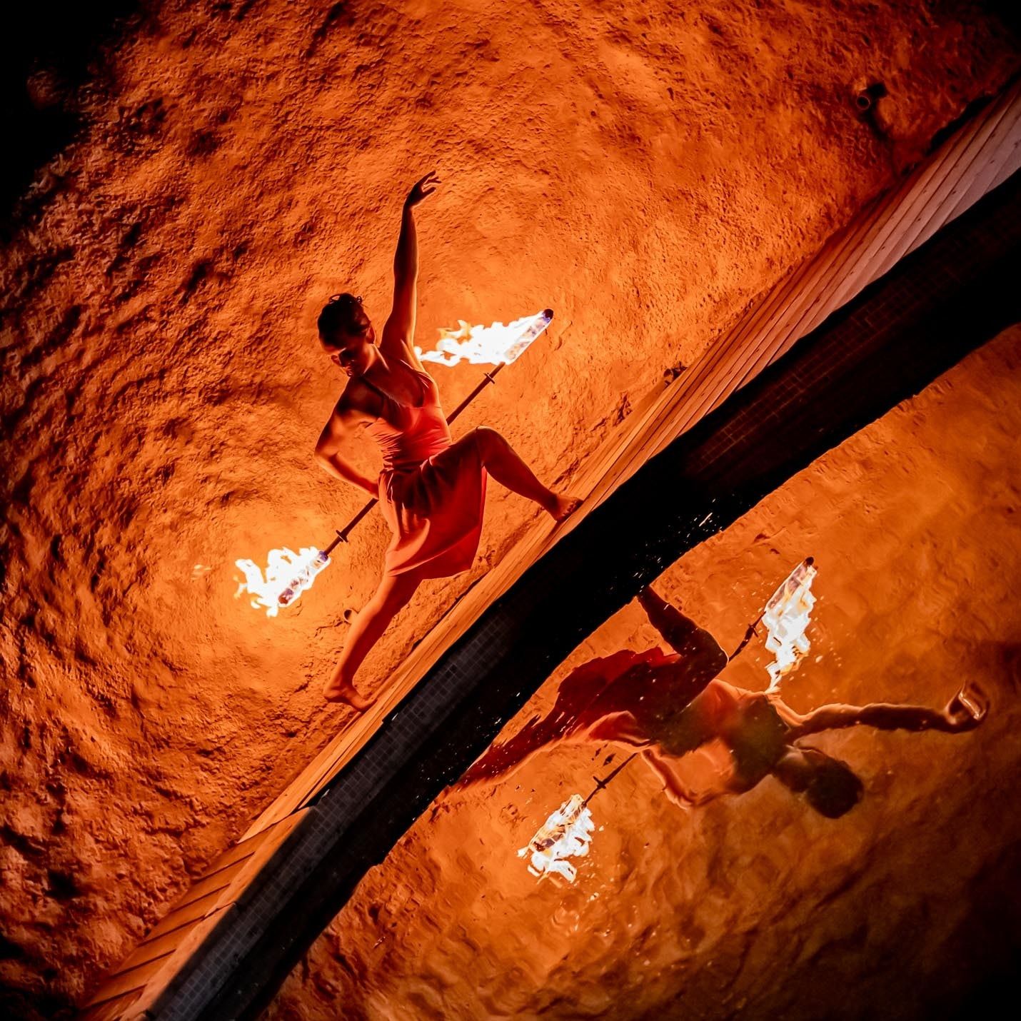 Vue de biais, la danseuse au bord d’un bassin, un bâton enflammé dans le dos ; son reflet danse dans l’eau, dans la lumière orange du feu.
