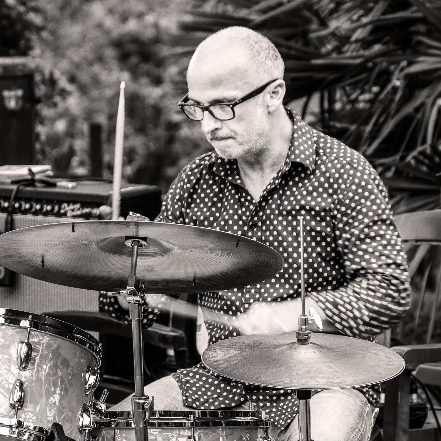 Un batteur du trio à ses cymbales, dans un jardin, en noir et blanc.