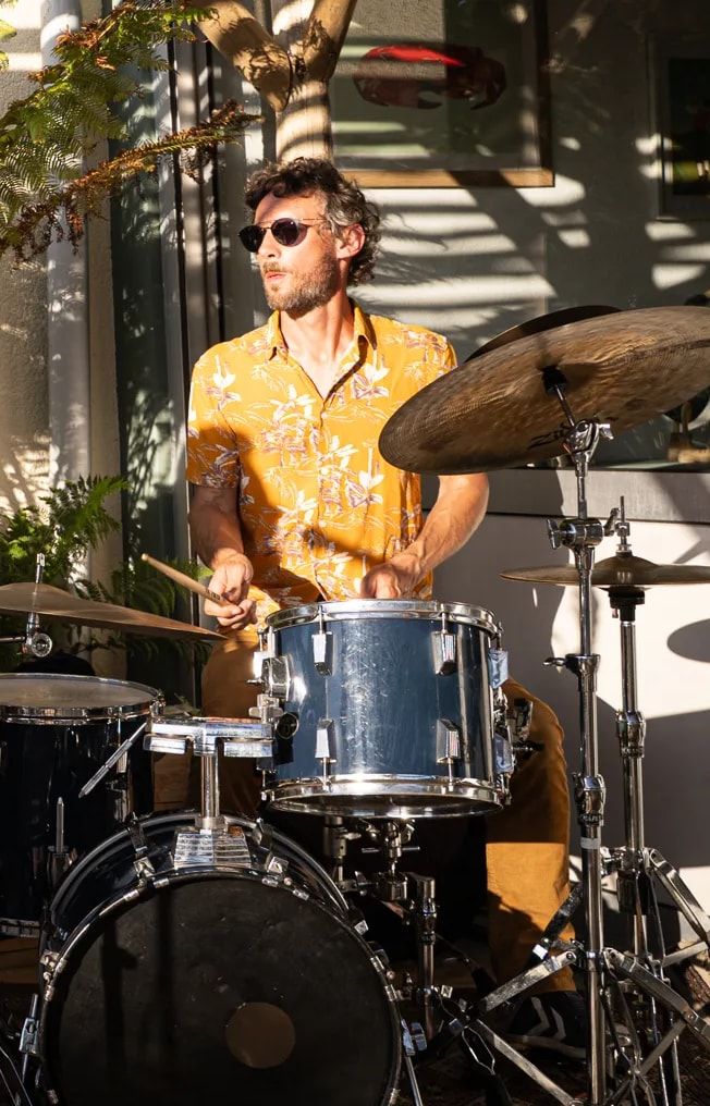 Le batteur, lunettes de soleil et chemise à fleurs, joue dans la lumière du soir.