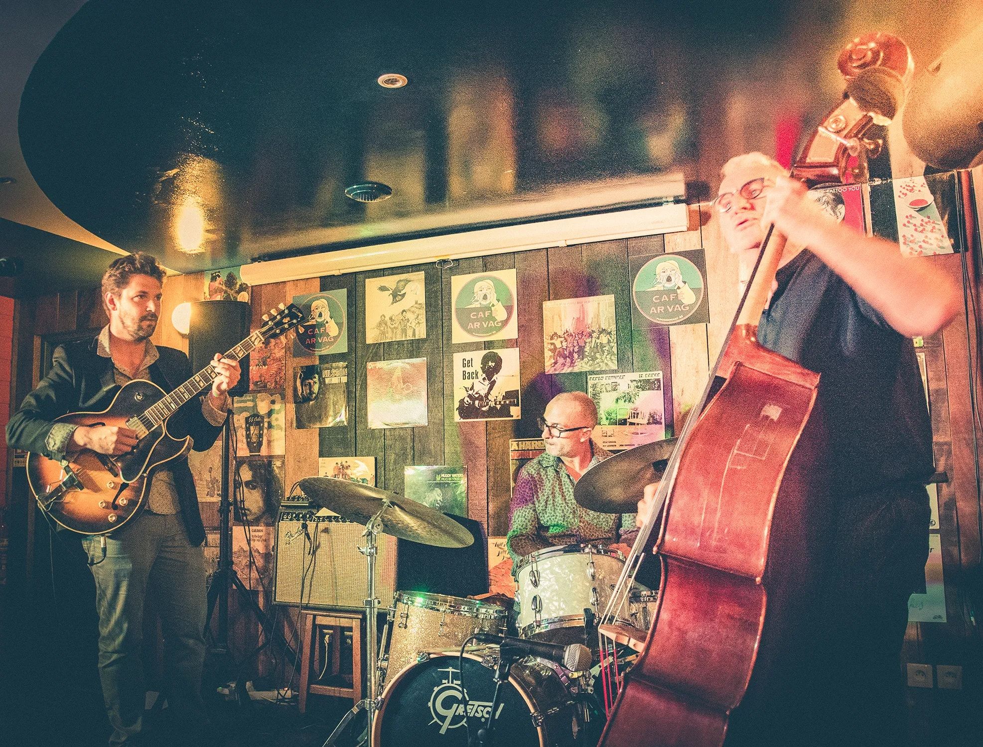 Dans un café-concert aux murs couverts de pochettes de disques, le trio joue : guitare, batterie et contrebasse.