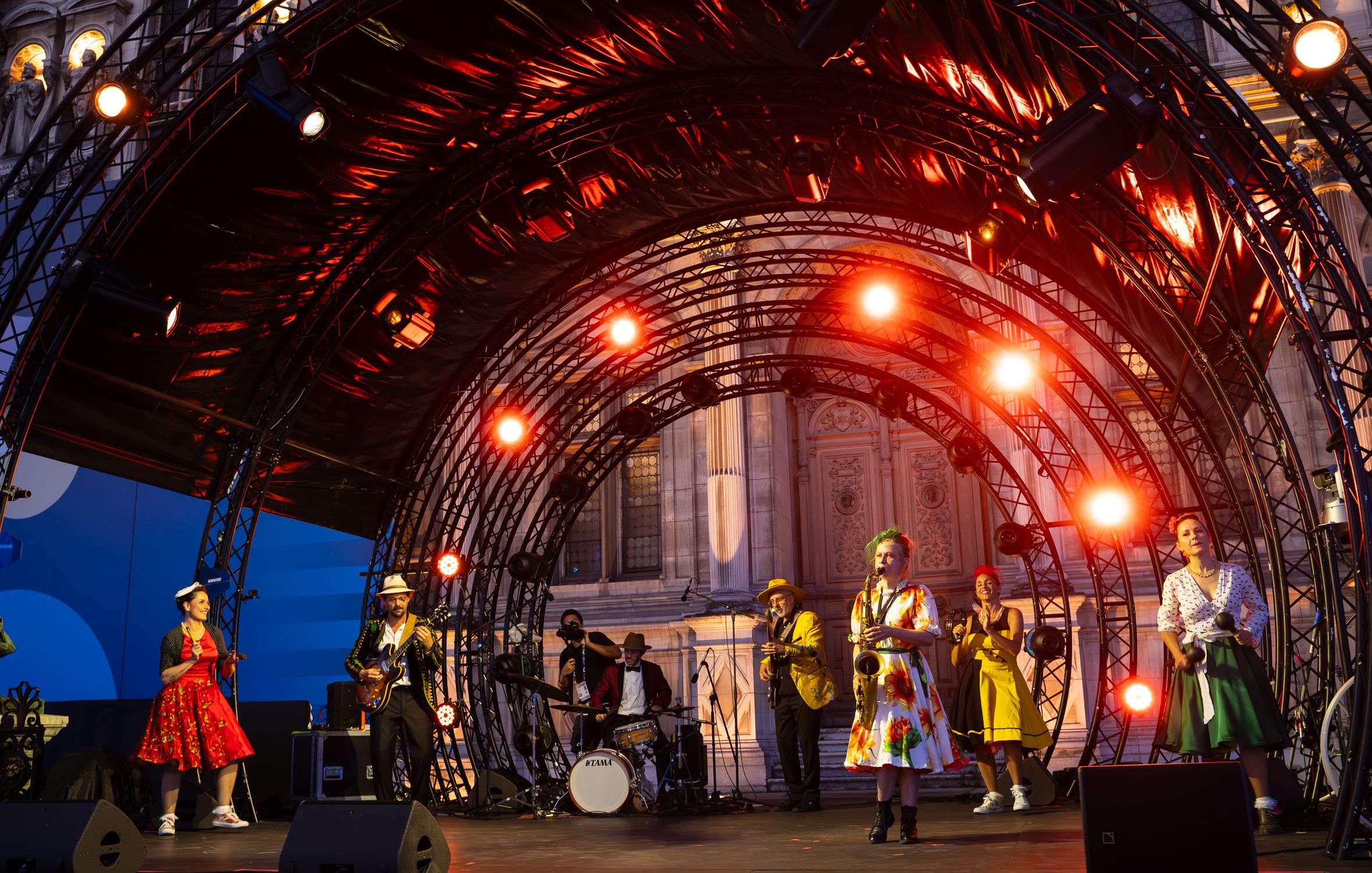 Les musiciens et danseuses de Soulful People sur une grande scène en plein air, sous une arche de projecteurs rouges, devant l'Hôtel de Ville de Paris.