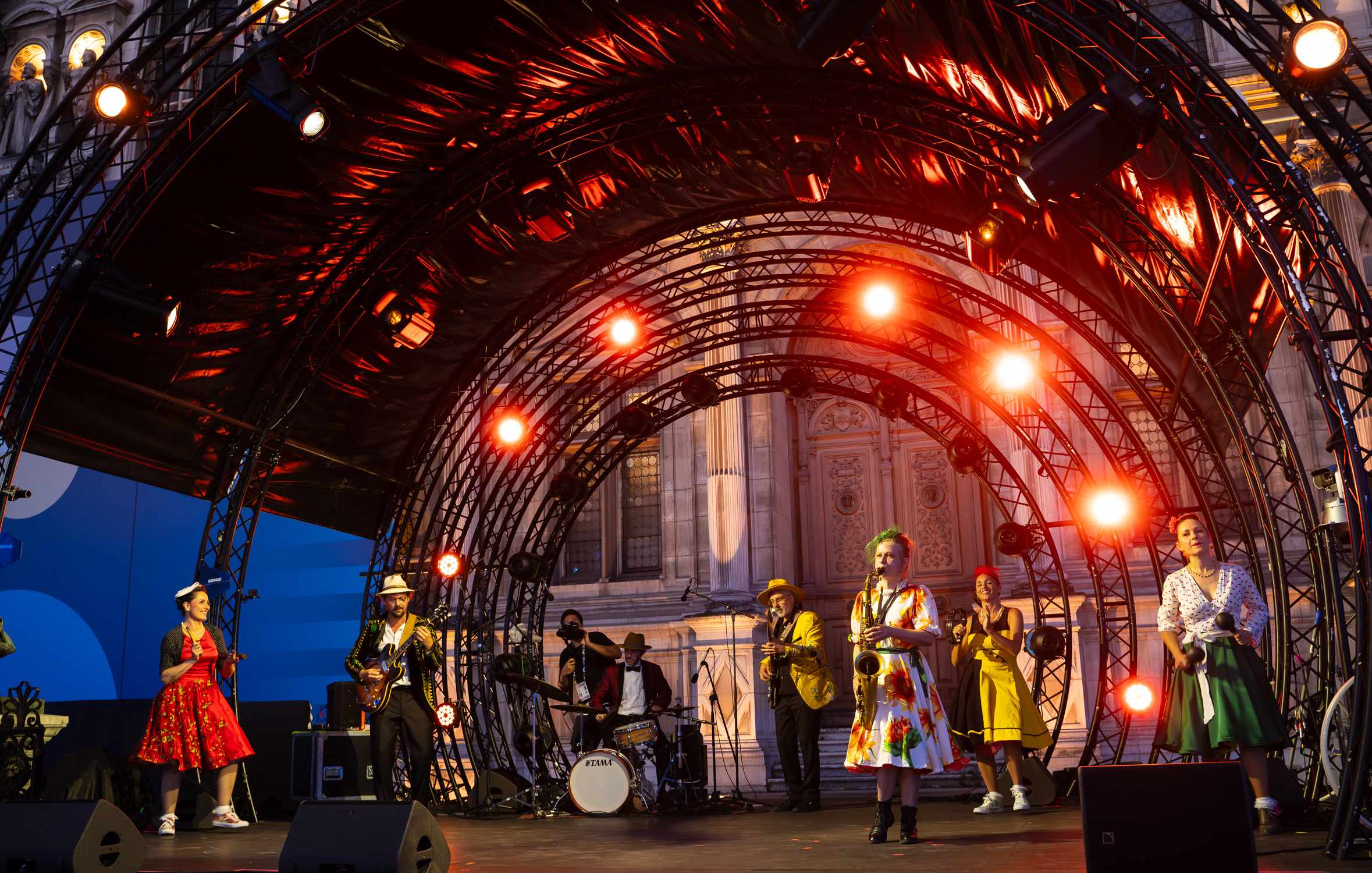 Les musiciens et danseuses de Soulful People sur une grande scène en plein air, sous une arche de projecteurs rouges, devant l'Hôtel de Ville de Paris.