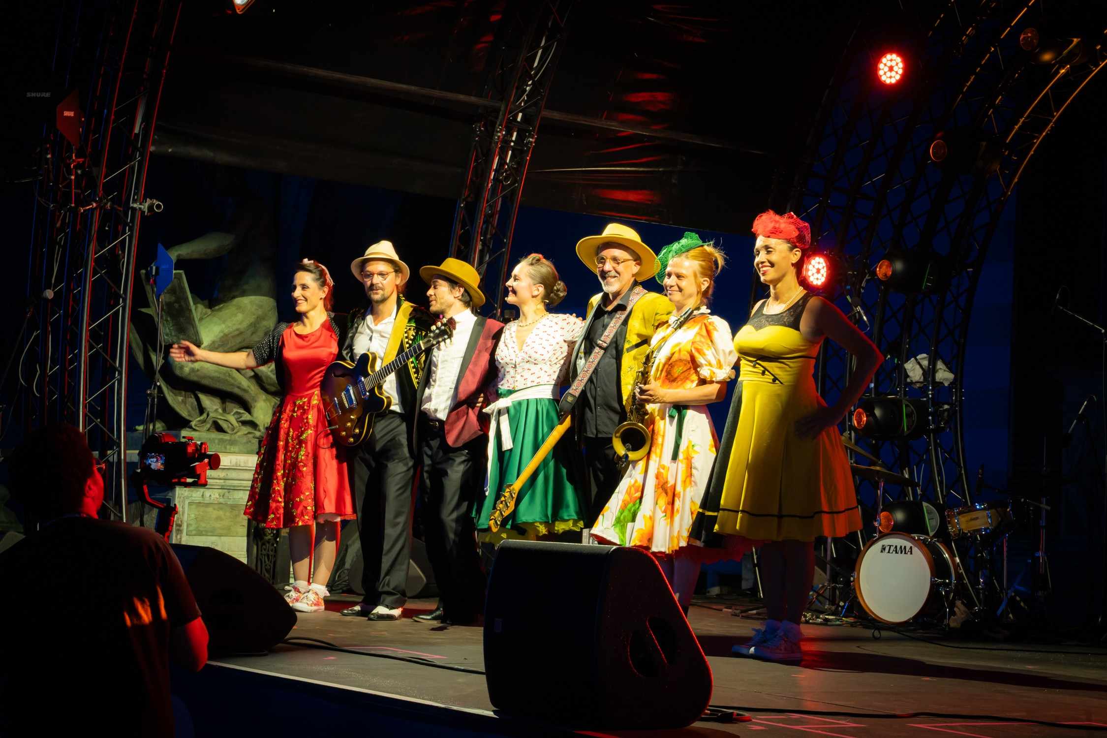 Les musiciens et danseuses de Streetside saluent sur scène en costumes colorés, chapeaux de paille et robes à fleurs, sous les projecteurs.