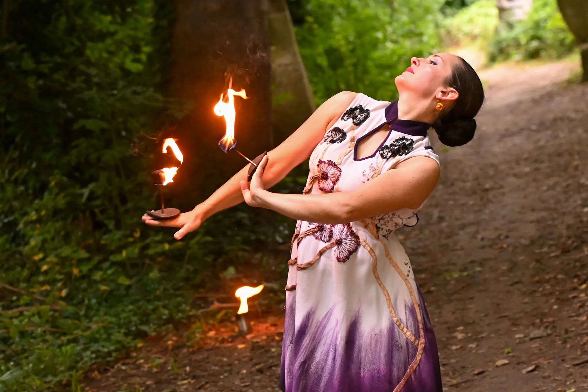 En forêt, de jour, la danseuse cambrée, le visage vers le ciel, des flammes au bout des doigts.