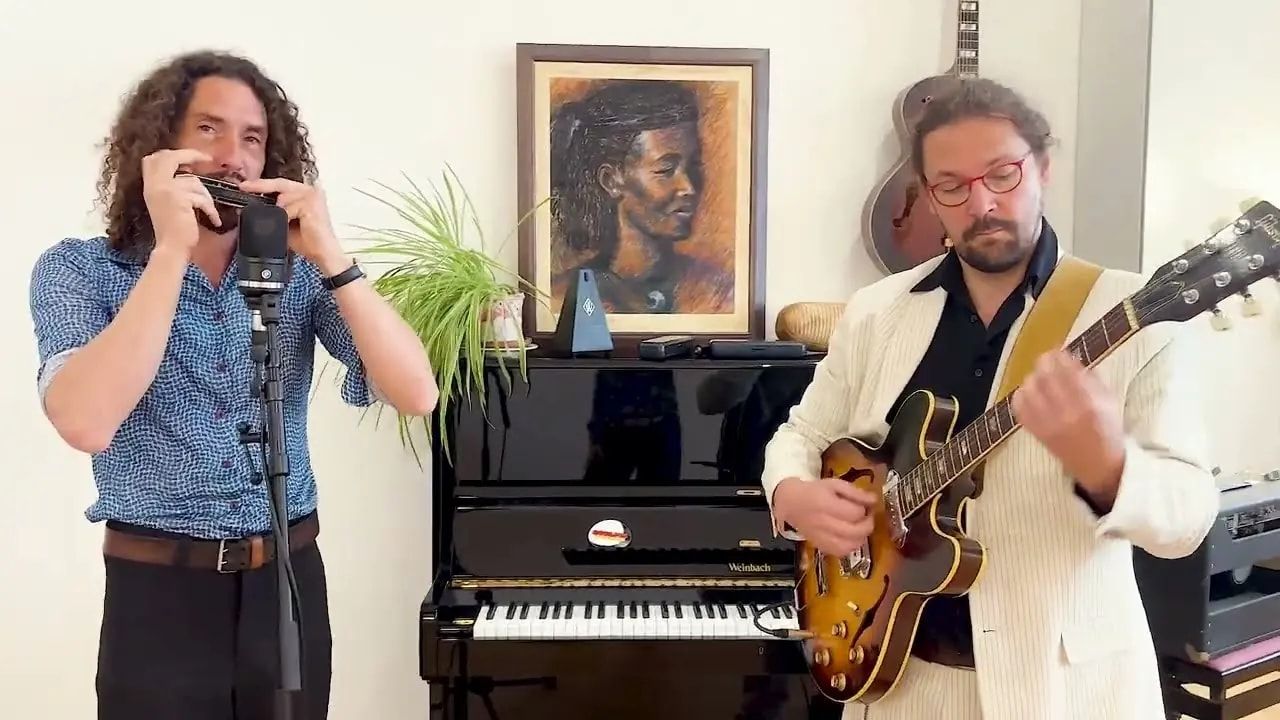 Dans un salon, devant un piano droit, Thomas Laurent joue de l’harmonica au micro et Mathieu Crochemore de la guitare.
