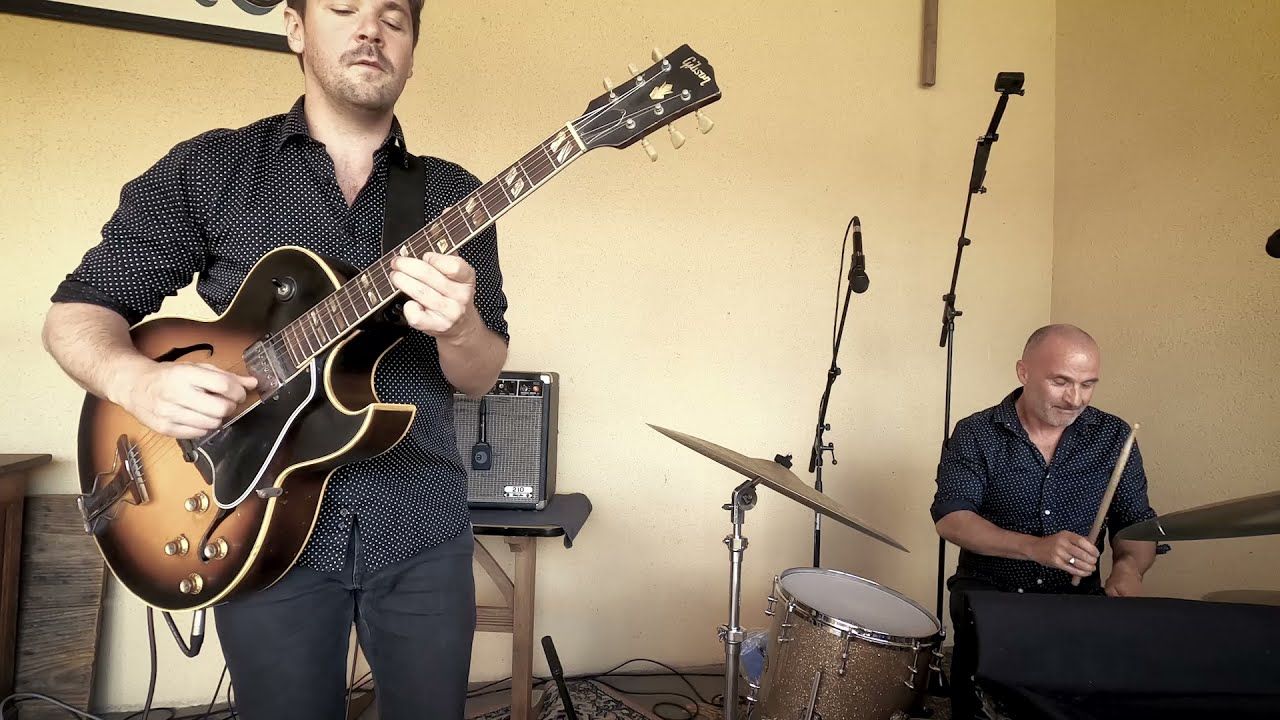 Le guitariste joue debout sur une guitare demi-caisse ; derrière lui, le batteur à ses cymbales.