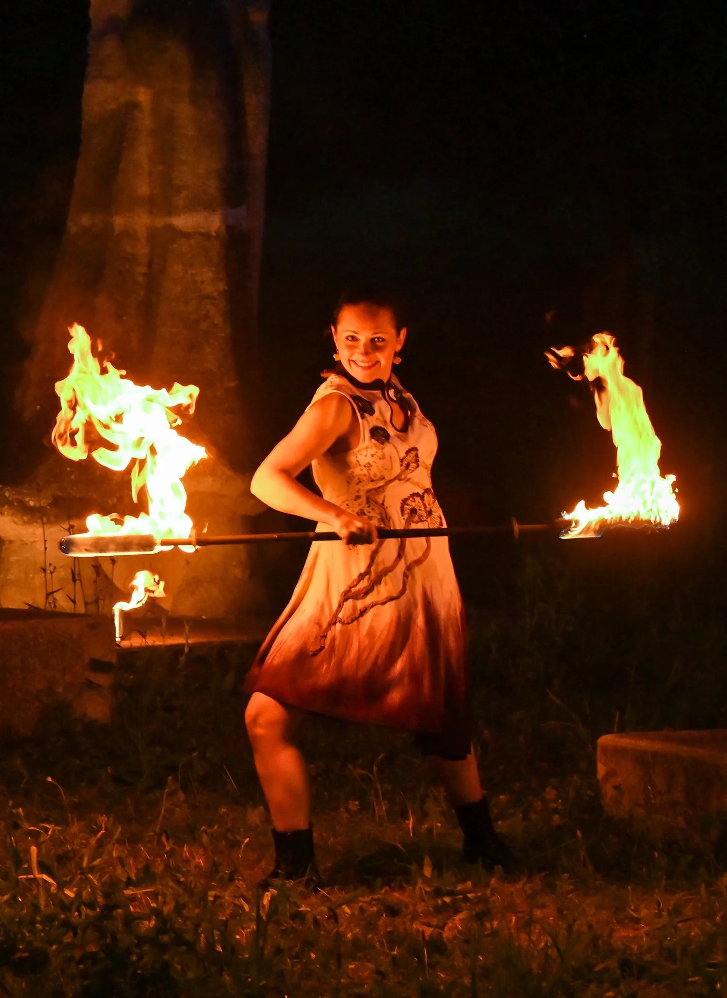 La nuit, en forêt, la danseuse tient un bâton enflammé à ses deux bouts, deux autres flammes autour d’elle.