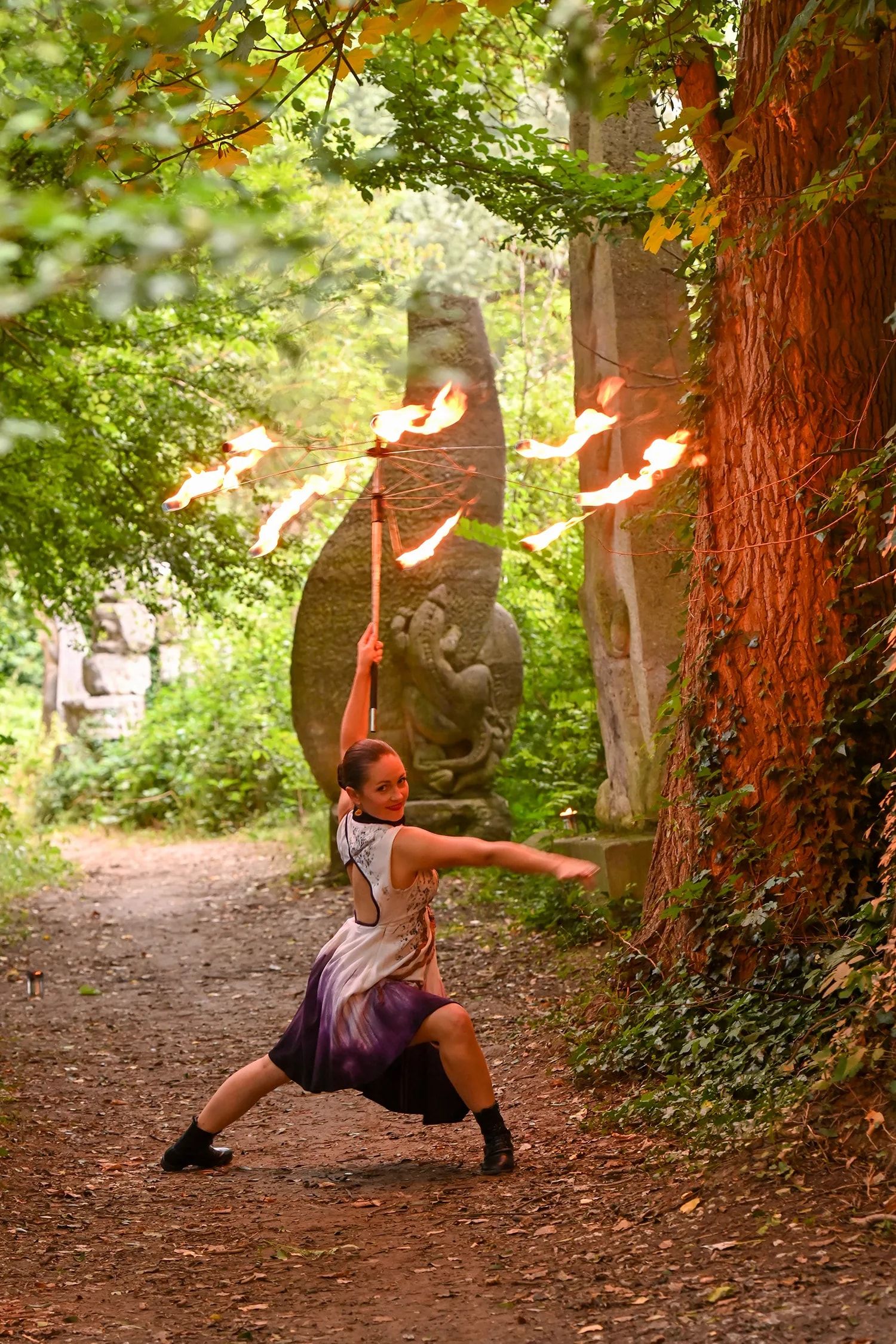 Dans un sentier, entre les arbres, la danseuse en fente lève un éventail de flammes au-dessus d’elle.