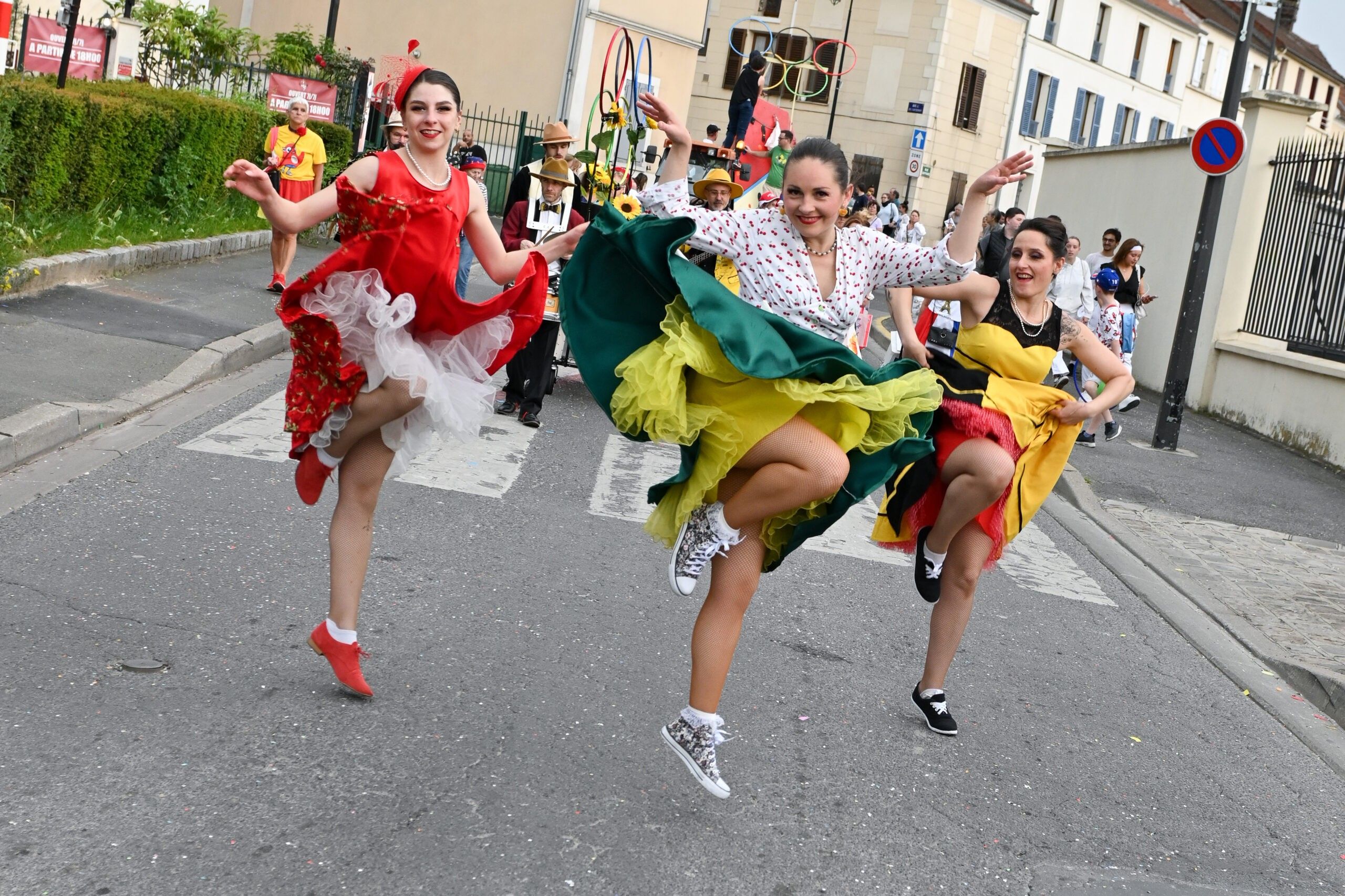 Trois danseuses de Streetside en pleine rue, robe rouge, jupe verte et robe jaune, la jambe levée et les jupons en l’air, devant les musiciens du cortège.