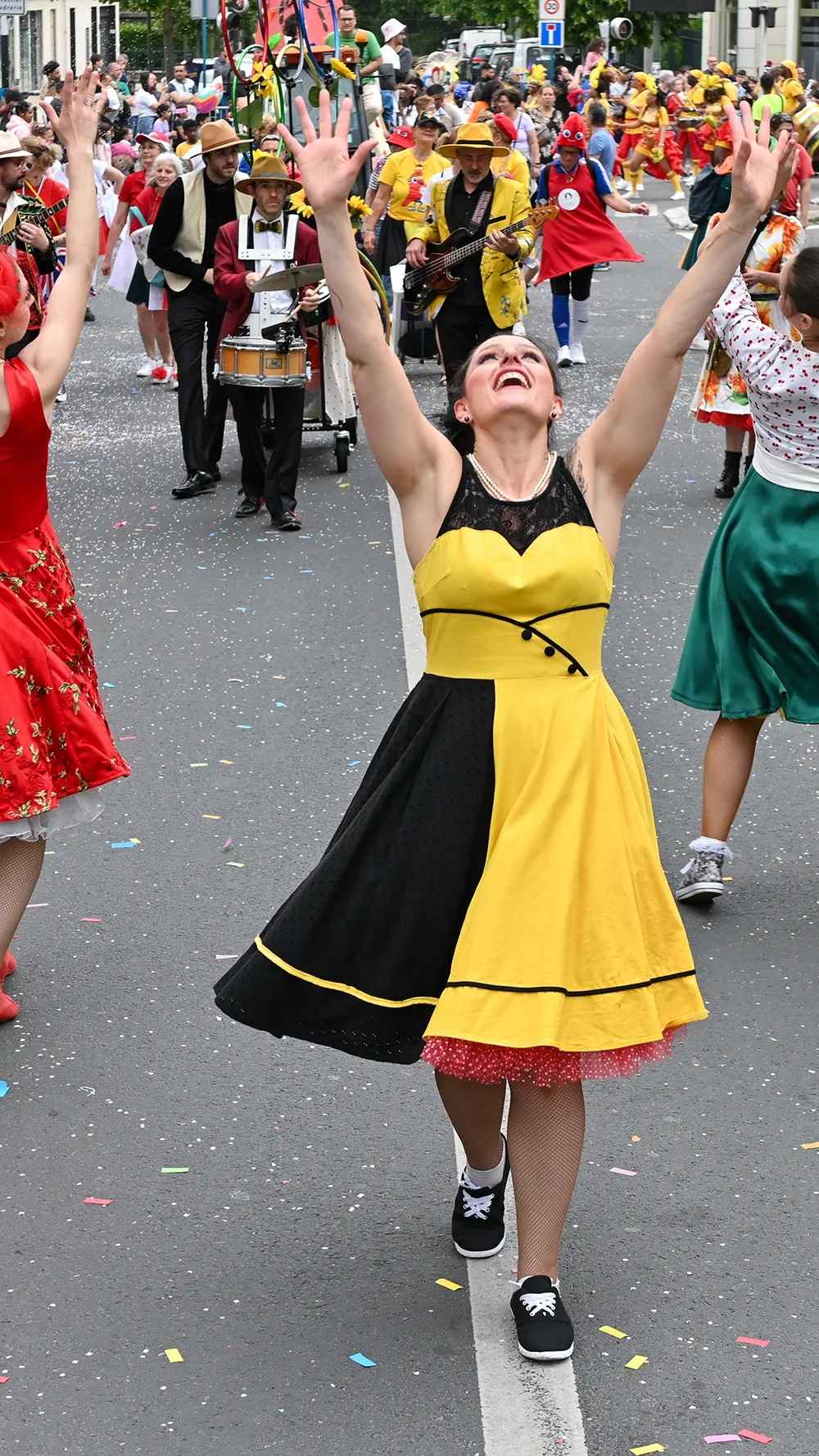 En plein jour, une danseuse de Streetside en robe jaune et noire avance dans la rue, les bras levés vers le ciel, devant le cortège.