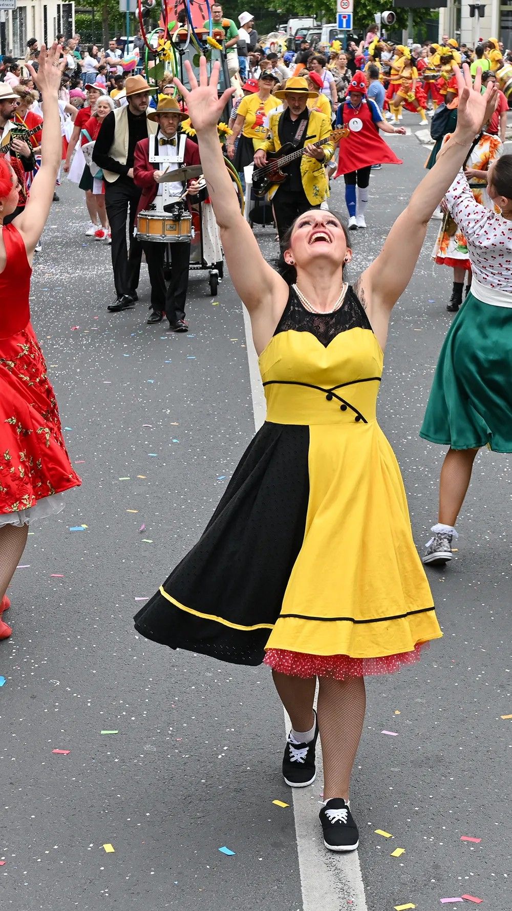 En plein jour, une danseuse de Streetside en robe jaune et noire avance dans la rue, les bras levés vers le ciel, devant le cortège.