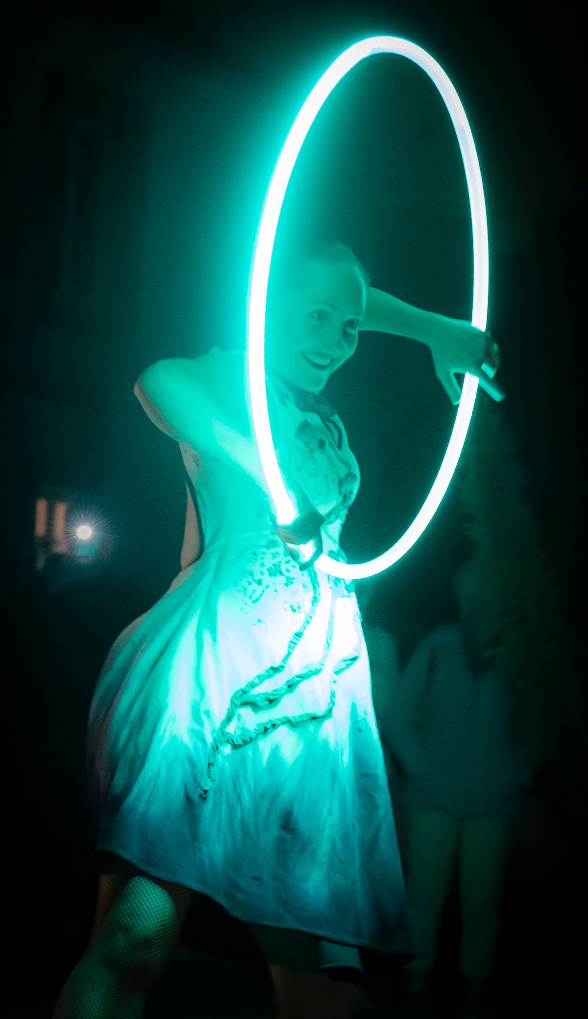 La nuit, une danseuse de Streetside en robe d’été tient son hoop lumineux, qui l’éclaire d’une lumière turquoise.