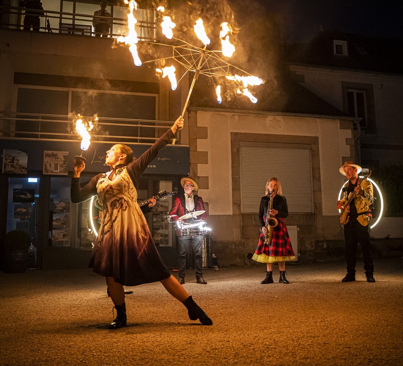 Sur une place, la nuit, une danseuse lève ses éventails de feu ; les musiciens jouent derrière elle.