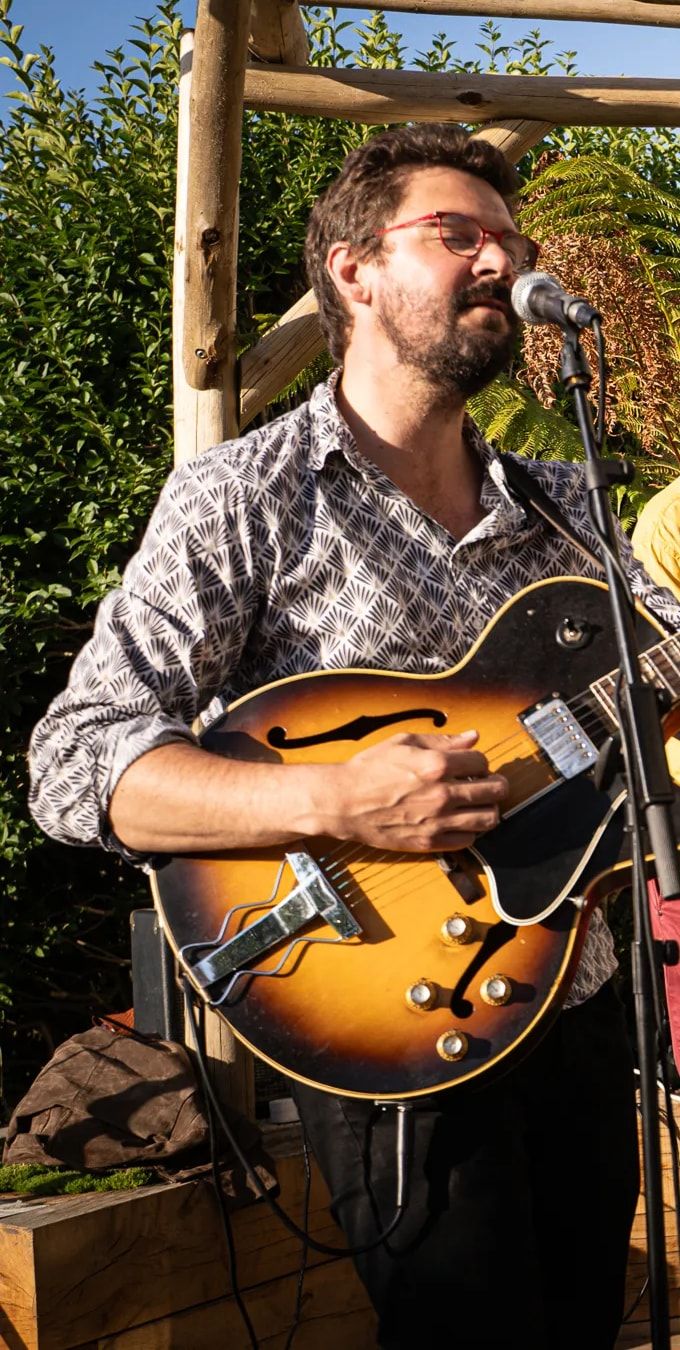 Le guitariste chante au micro, guitare demi-caisse en main, sous une pergola au soleil.