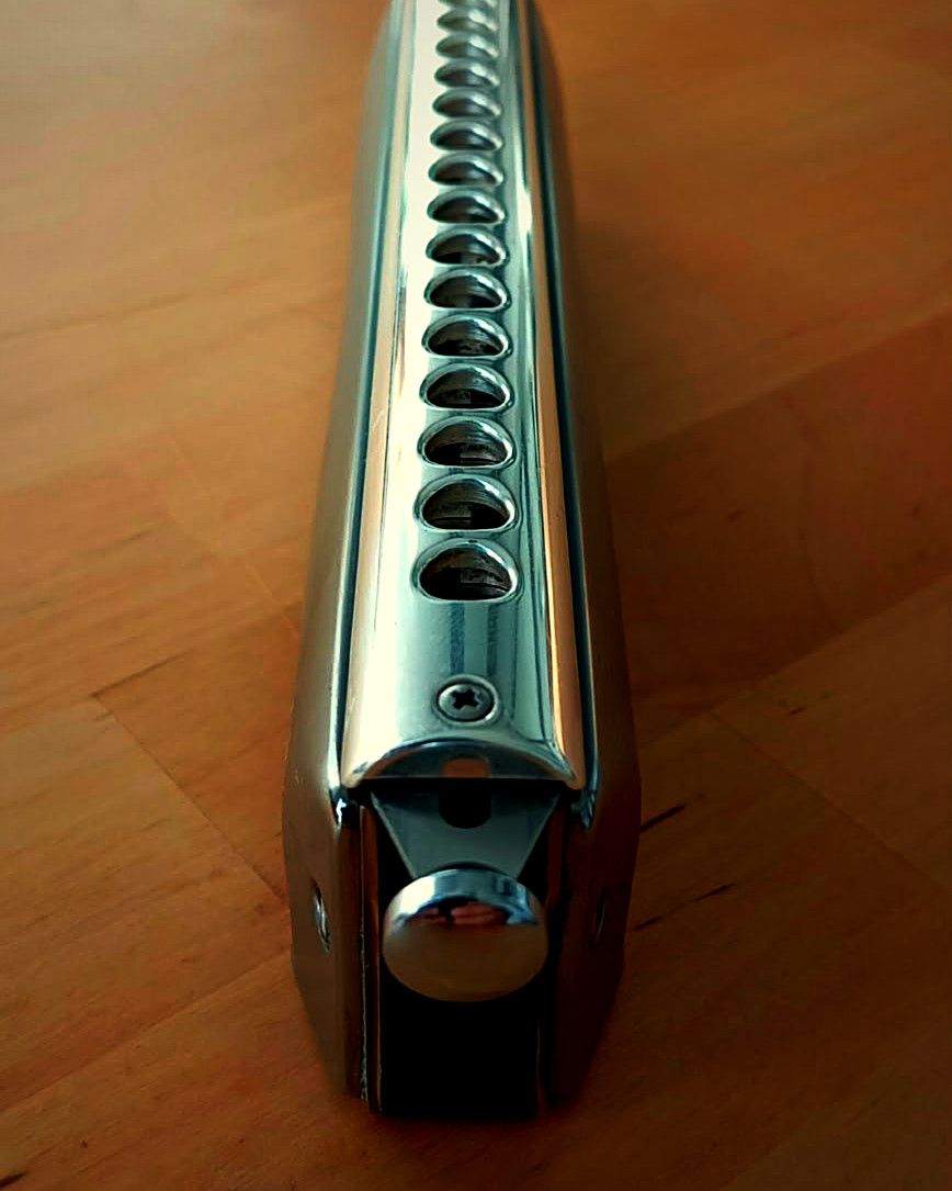 Un harmonica chromatique posé sur une table en bois, vu dans sa longueur : le bouton de la tirette au premier plan, puis les trous de l’embouchure chromée qui filent vers le fond.