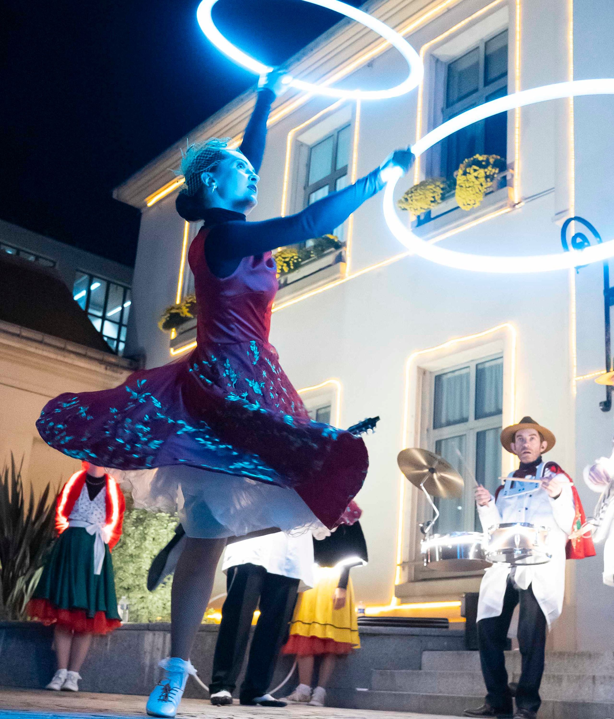 Le soir, une danseuse de Streetside en robe brodée tourne, le bras levé vers son hoop lumineux, devant une façade illuminée.