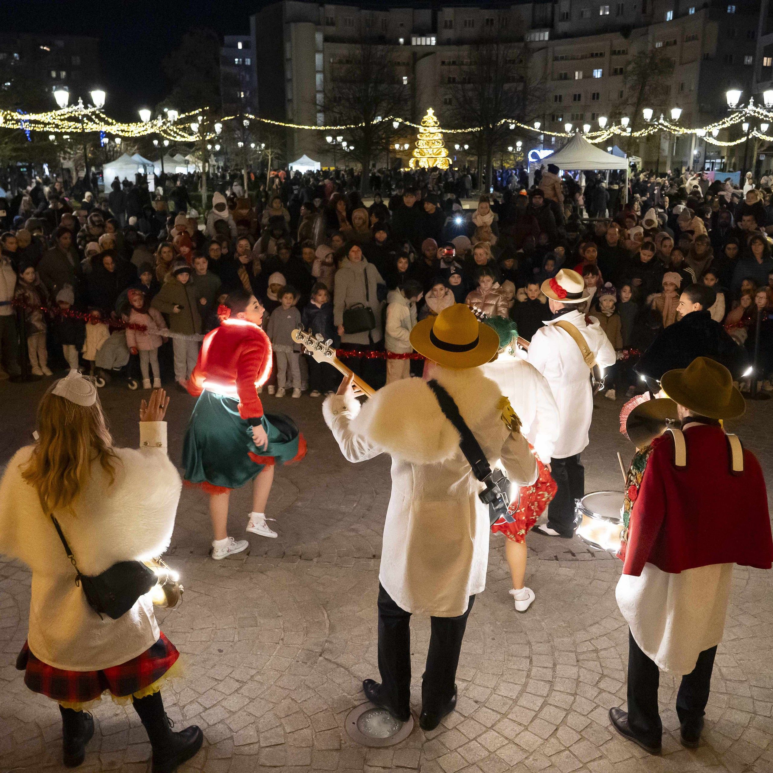 La nuit, sur une place illuminée pour Noël, les musiciens et les danseuses de Streetside au milieu du public.