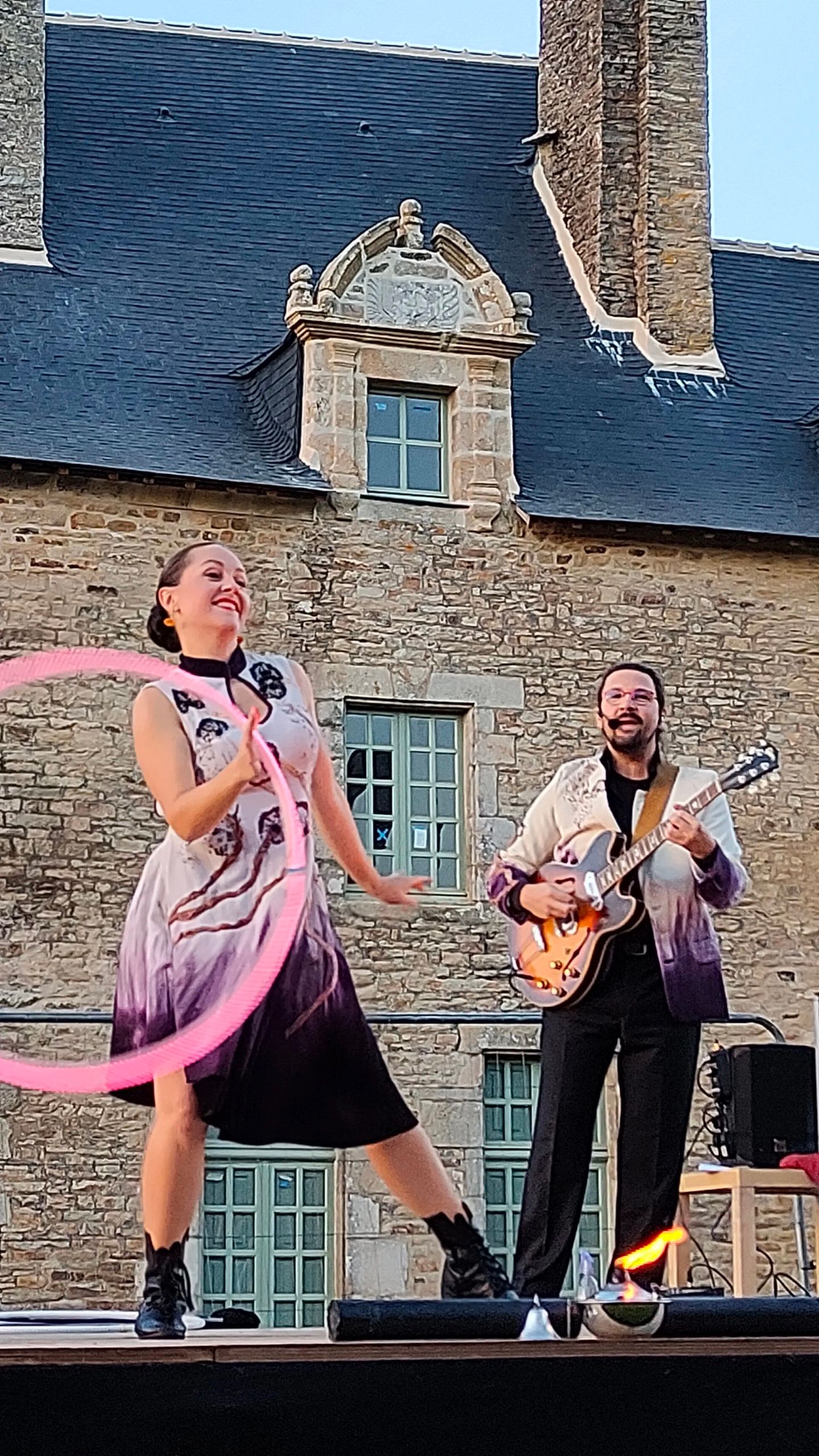 En plein jour, sur une scène devant un manoir, la danseuse fait tourner un hula hoop rose ; le guitariste joue en souriant.