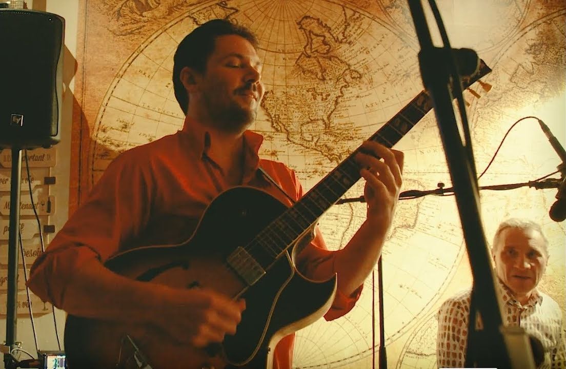 Le guitariste du trio joue les yeux fermés, en chemise orange, devant une carte ancienne du monde.