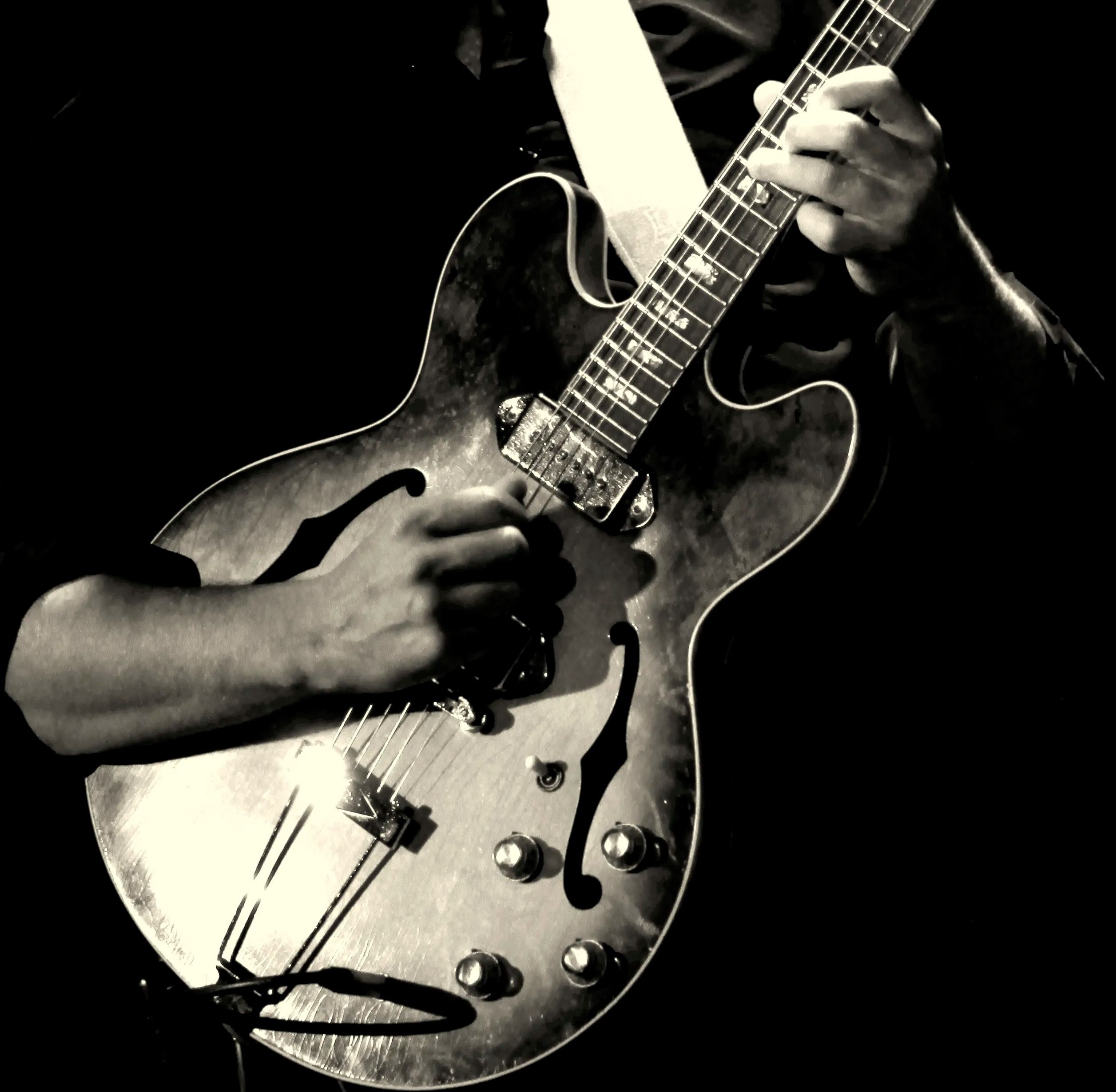En noir et blanc, deux mains sur une guitare demi-caisse : l’une sur le manche, l’autre sur les cordes.