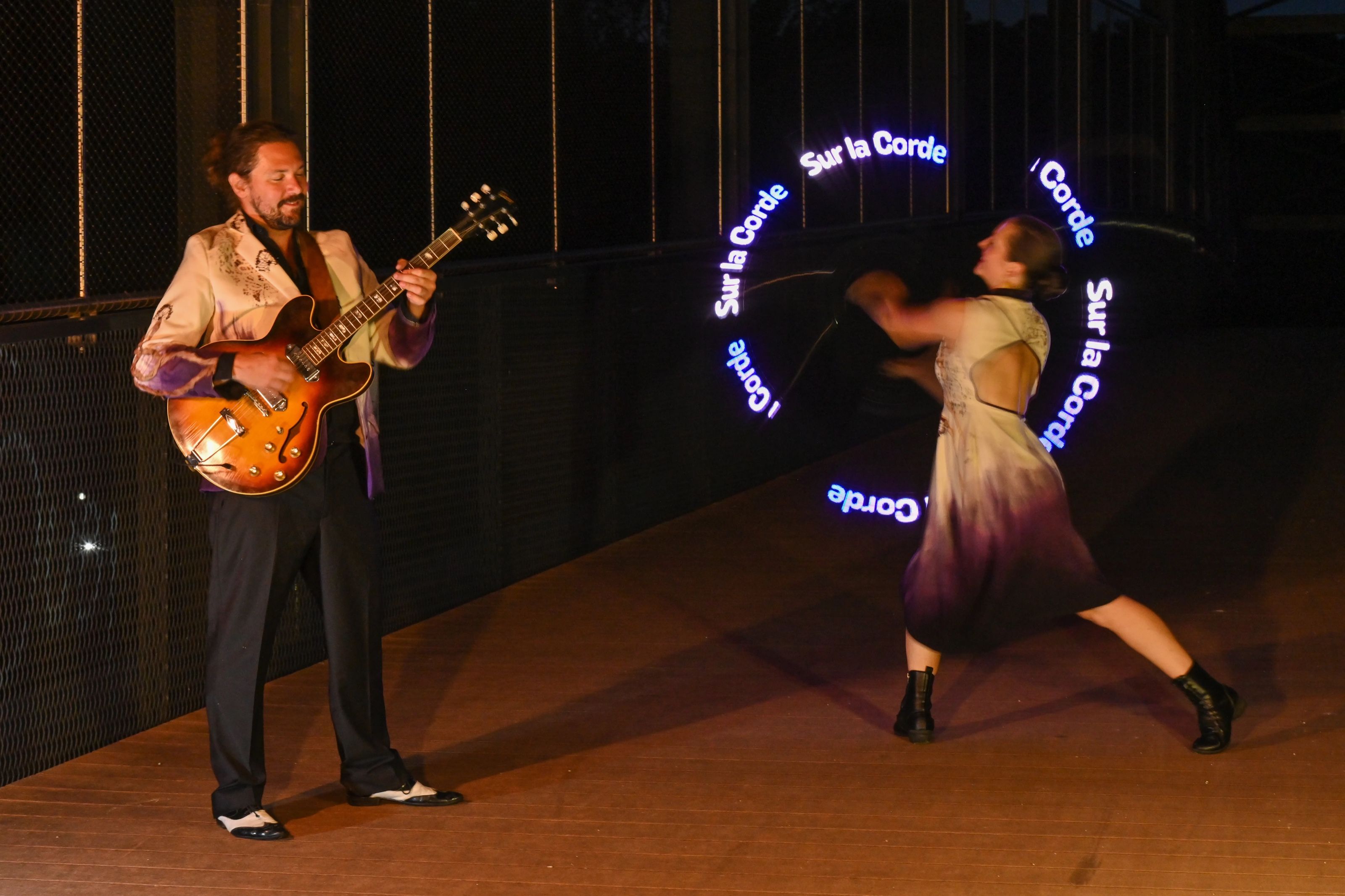 La nuit, un guitariste joue face à une danseuse qui fait tourner un cercle de lumière où s’écrit « Sur la Corde ».