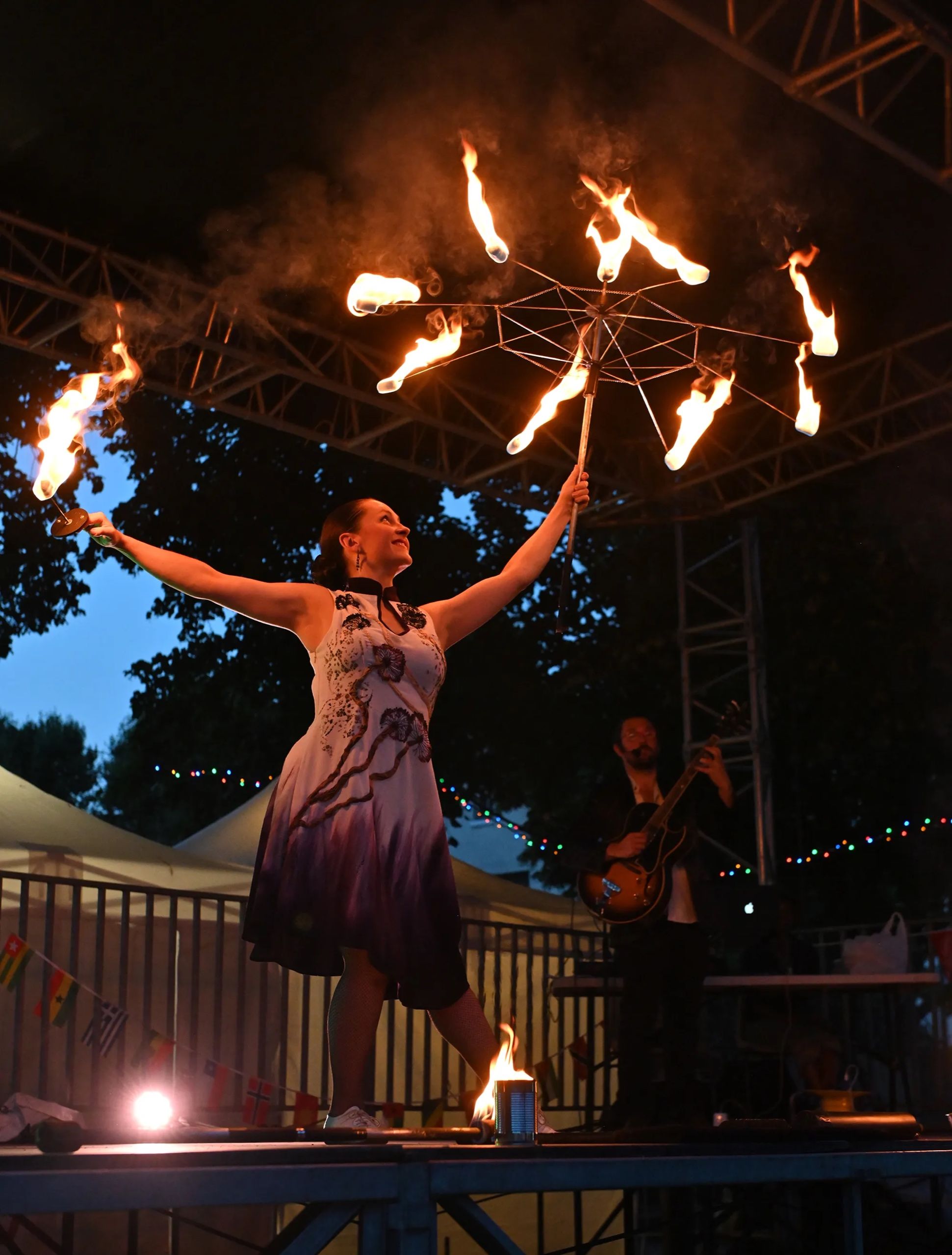 Sur scène, à la tombée du jour, la danseuse lève une roue de flammes au-dessus d’elle ; le guitariste joue derrière.