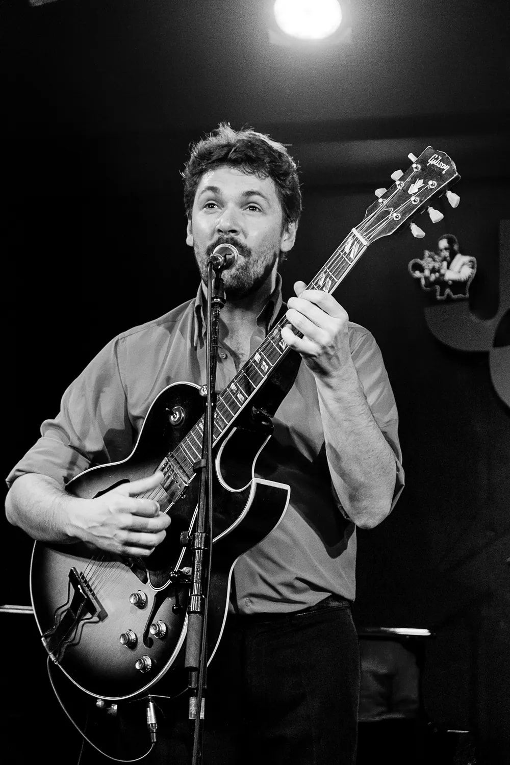 En noir et blanc, Mathieu Crochemore chante au micro en jouant de la guitare, sous un projecteur.