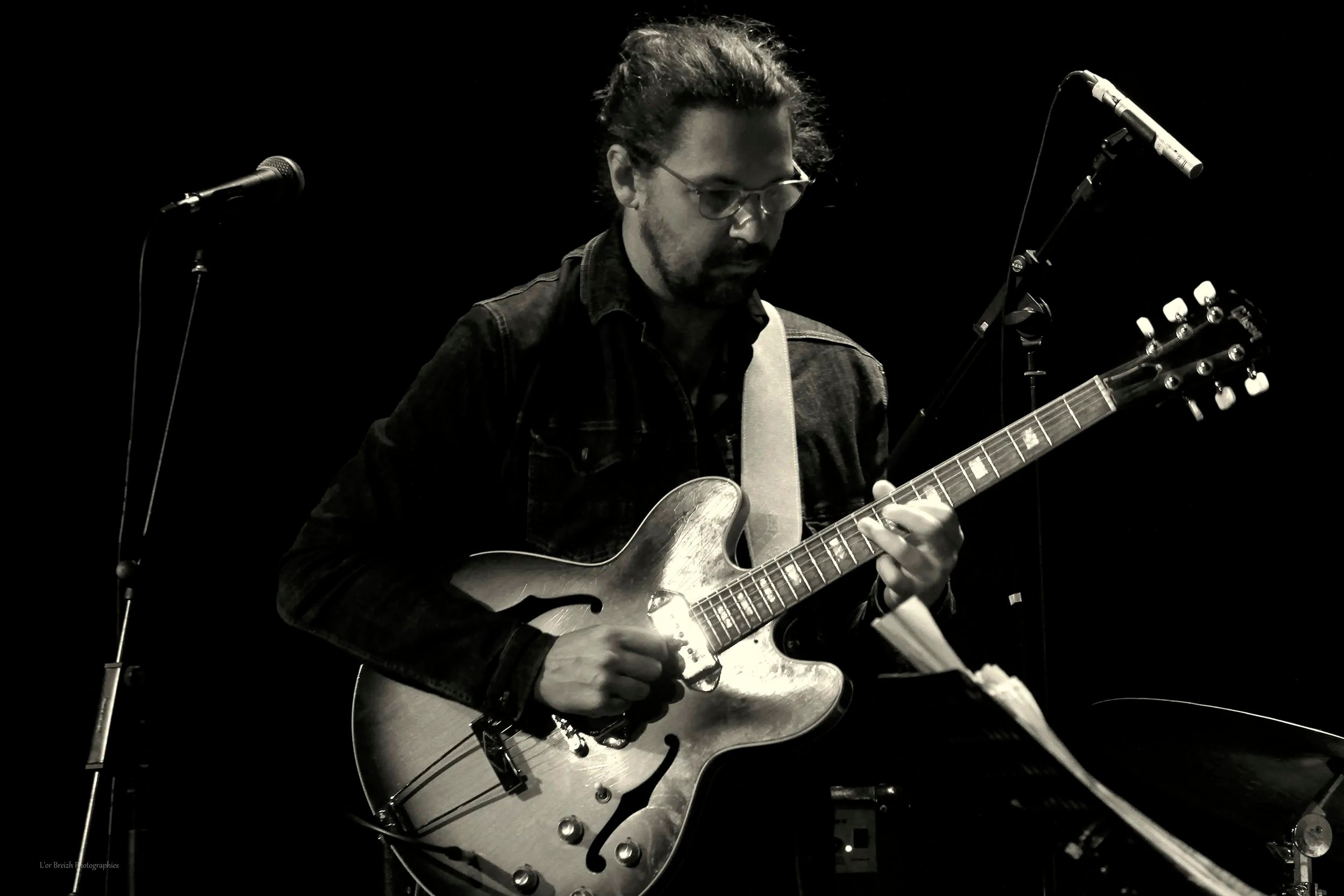 Sur scène, en noir et blanc, Mathieu Crochemore penché sur sa guitare demi-caisse, entre deux micros.
