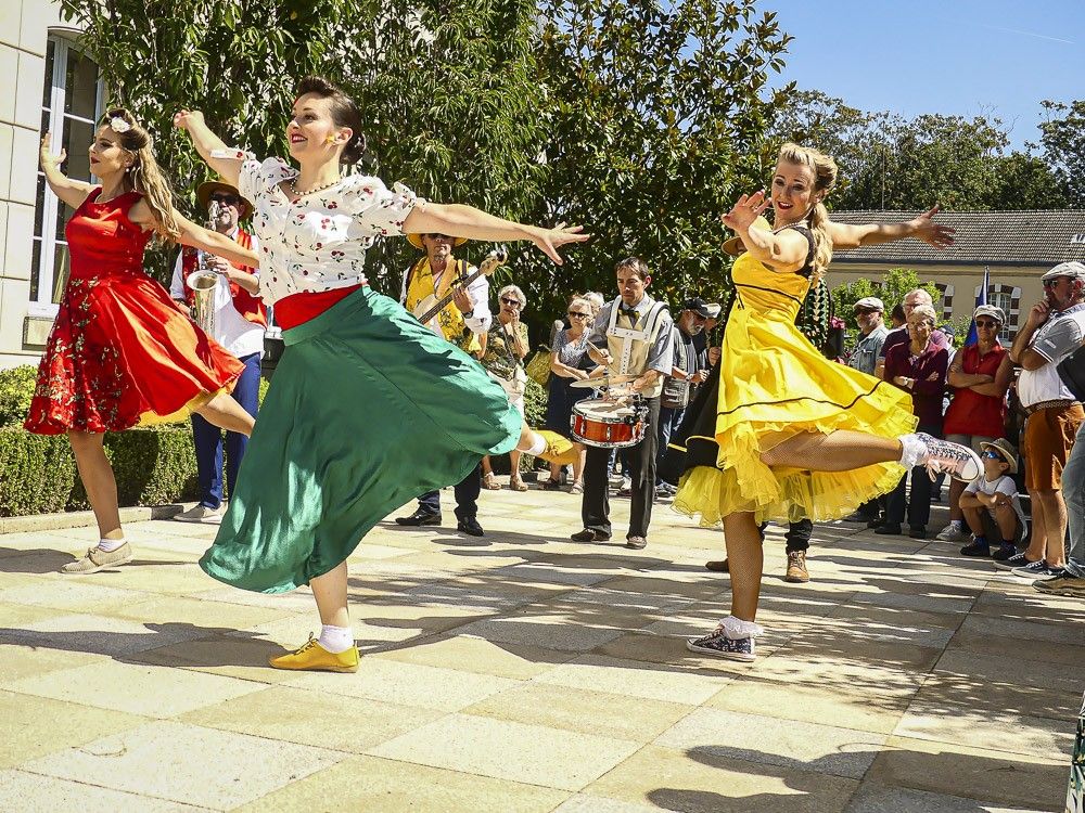 Sur une place ensoleillée, trois danseuses de Streetside en robes rouge, verte et jaune dansent devant les musiciens et le public assis.