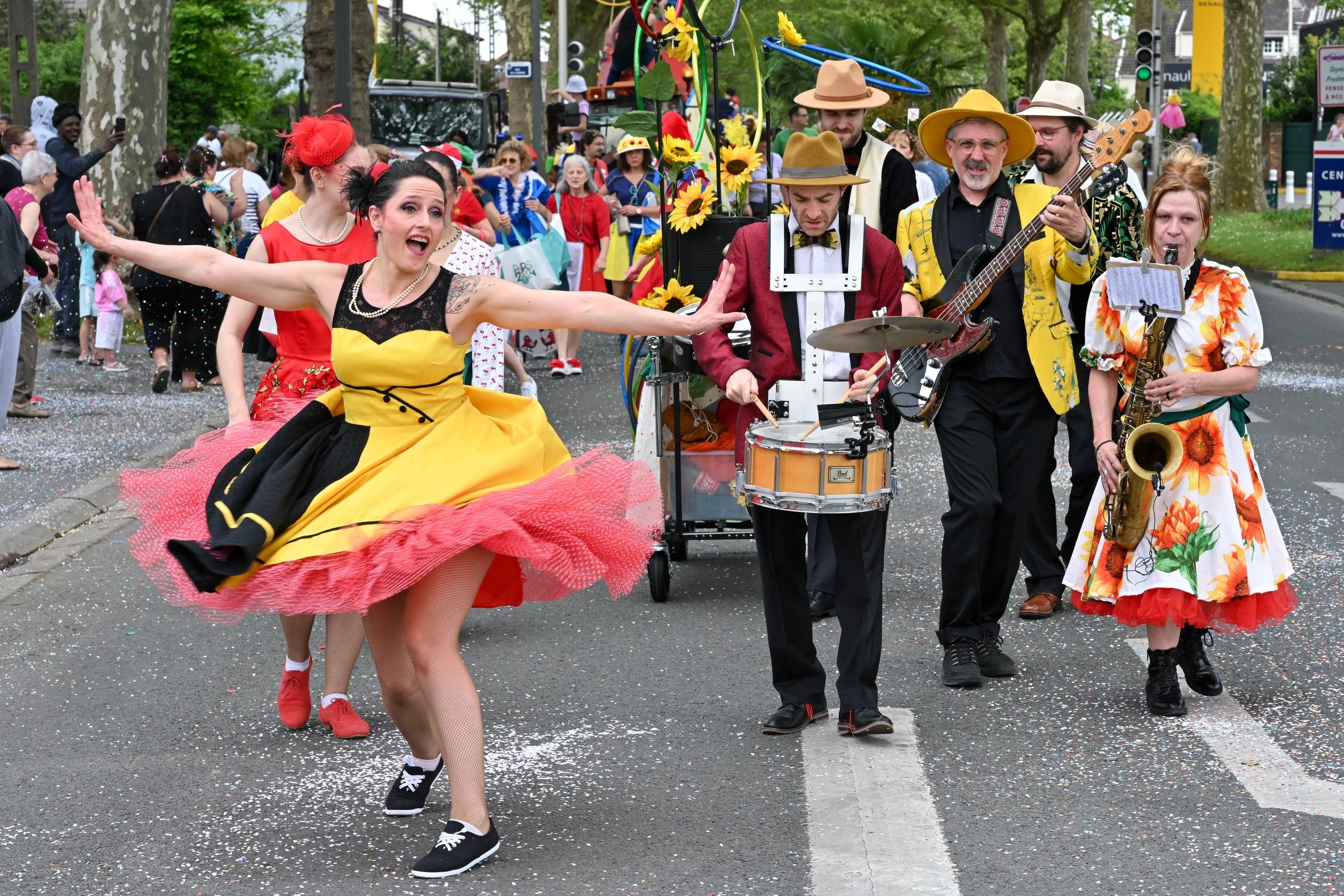 En pleine rue, une danseuse en robe jaune et jupon rose ouvre grand les bras devant les musiciens du cortège : caisse claire, basse et saxophone, chapeaux et vestes de couleur.