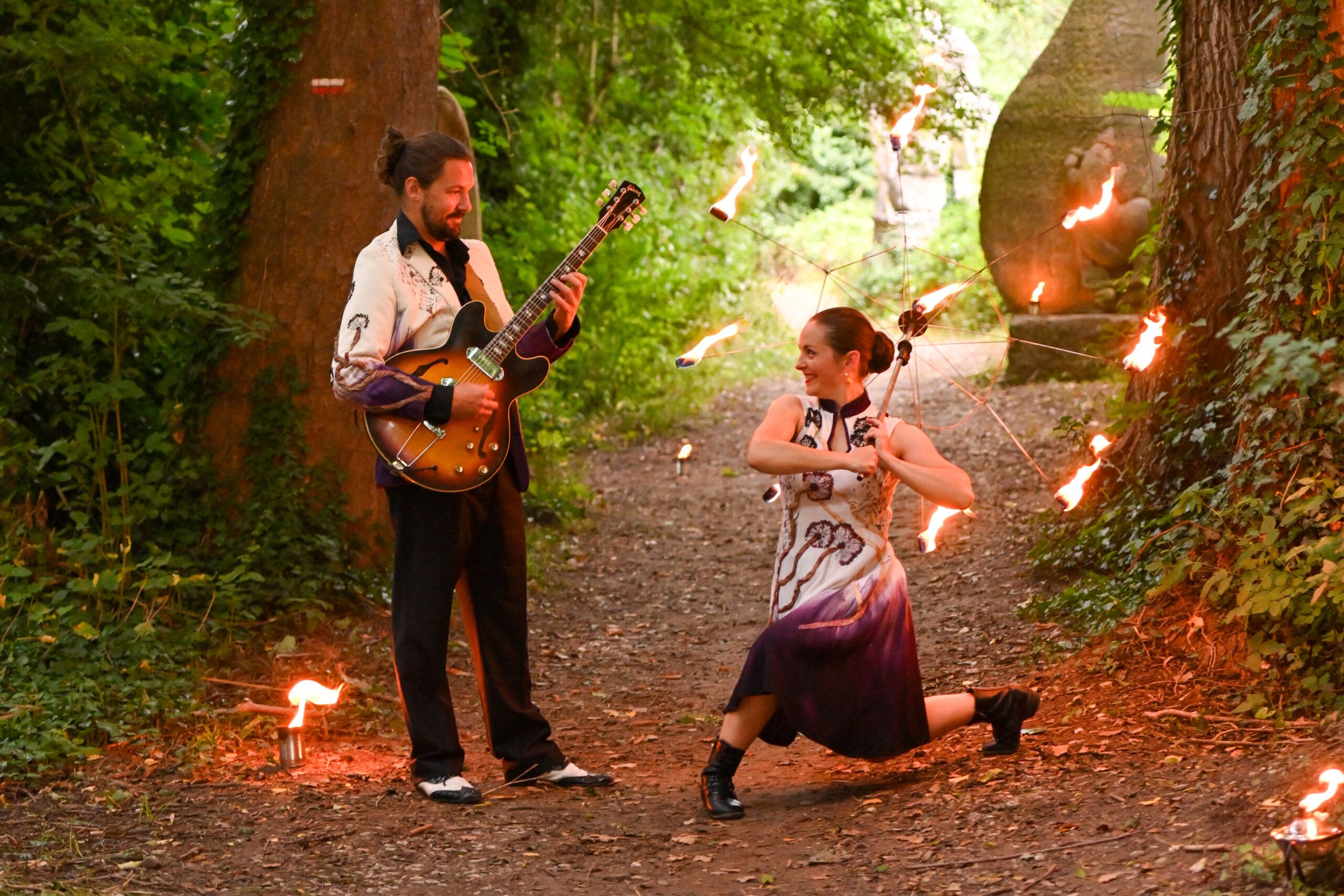 Dans un sentier, en plein jour, le guitariste joue tourné vers la danseuse, accroupie, un éventail de feu à la main.
