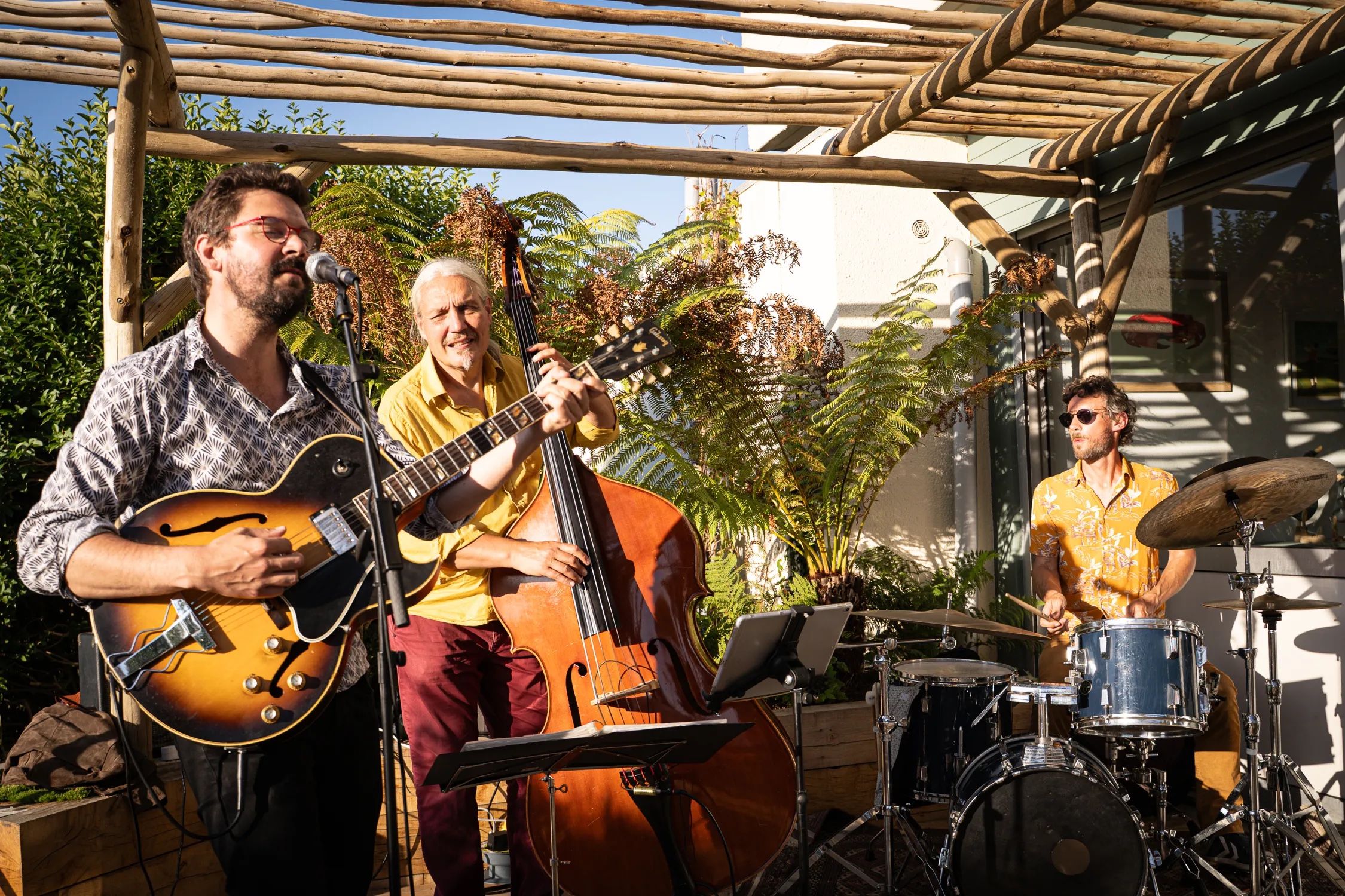 Le Soulful People Trio en concert sous une pergola, au soleil : le contrebassiste en chemise jaune, la batterie derrière lui, entre les fougères.