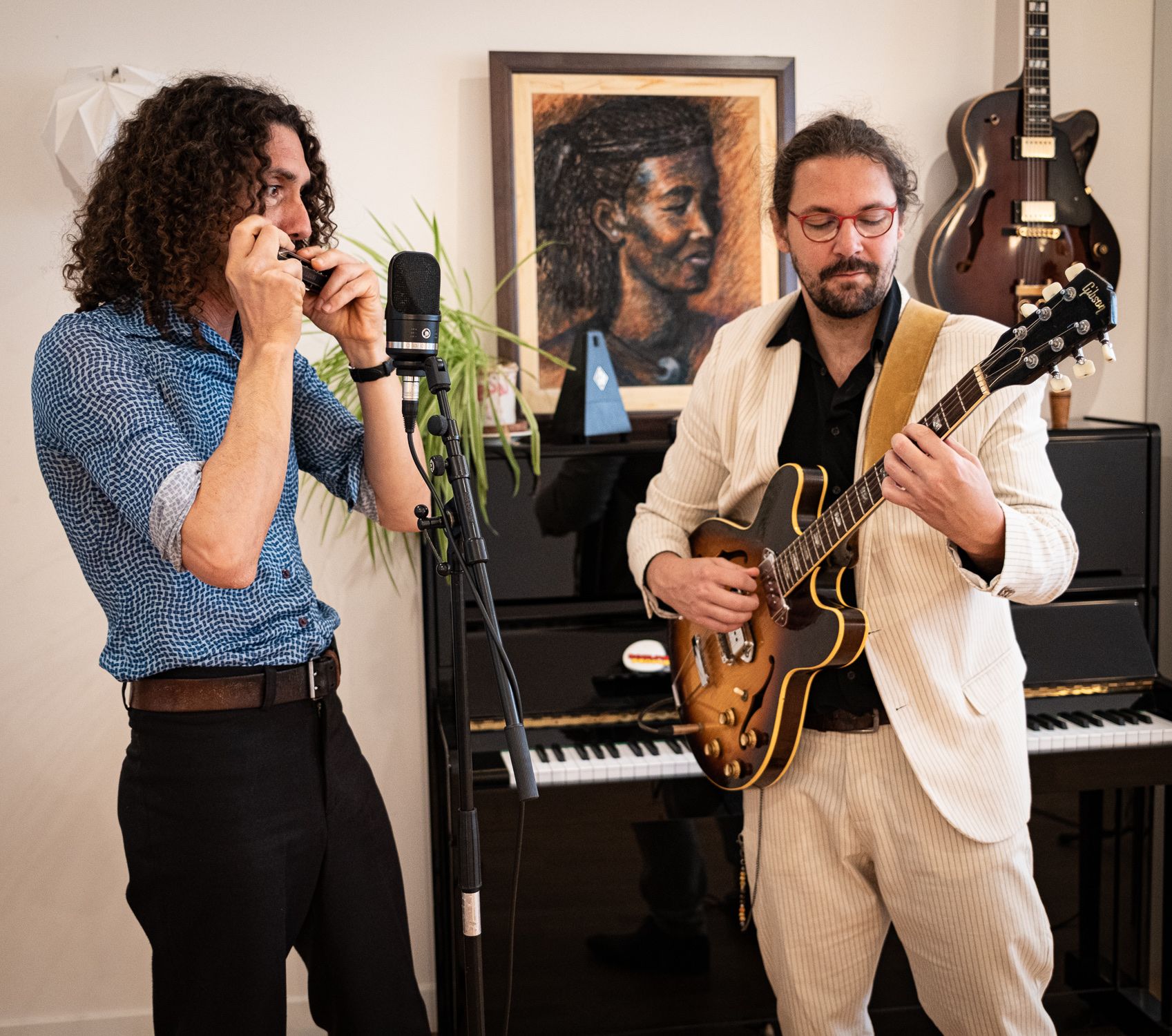 Dans un salon, Thomas Laurent, en chemise bleue, joue de l’harmonica au micro ; face à lui, Mathieu Crochemore, en costume clair, joue d’une guitare demi-caisse. Derrière eux, un piano droit et un portrait au pastel.