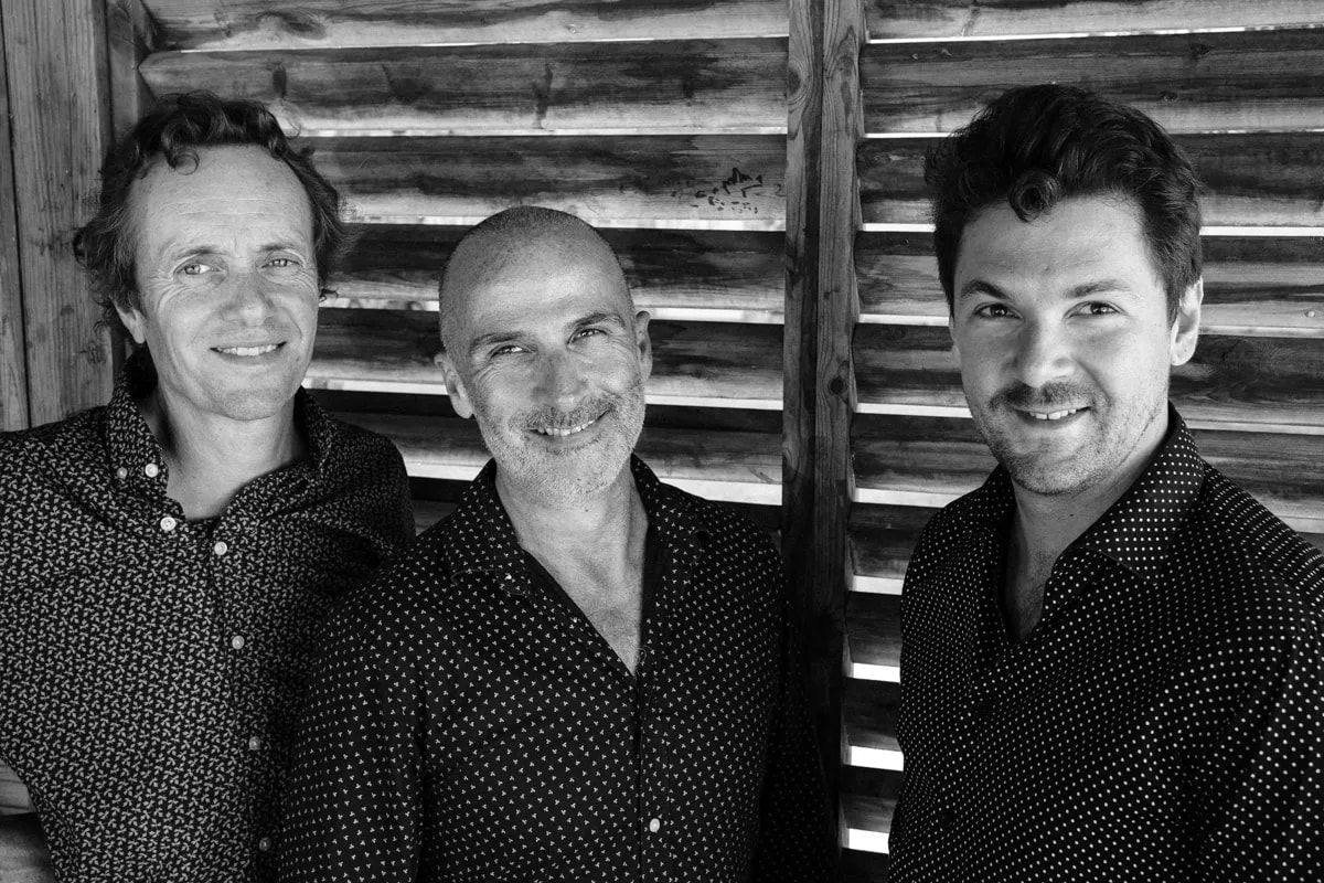 Les trois musiciens d’Ease Back Trio, souriants, en noir et blanc, devant des persiennes de bois.