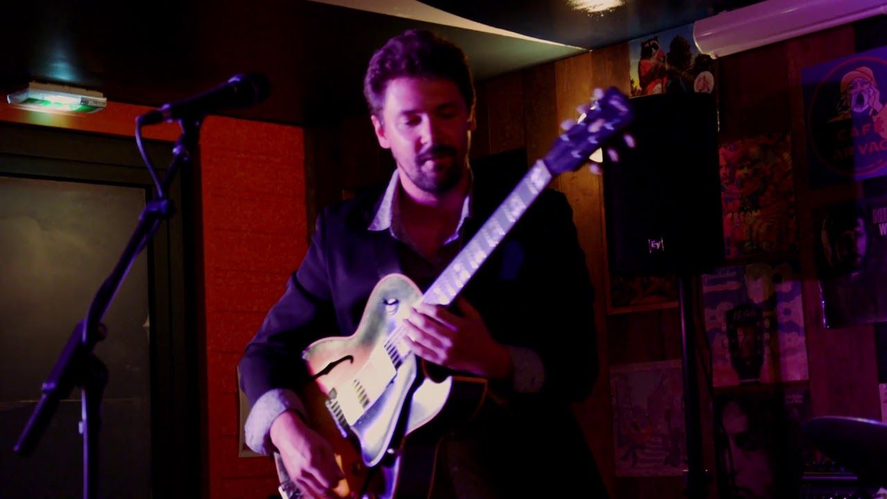 Dans un club, sous une lumière violette, le guitariste du trio joue, penché sur sa guitare.