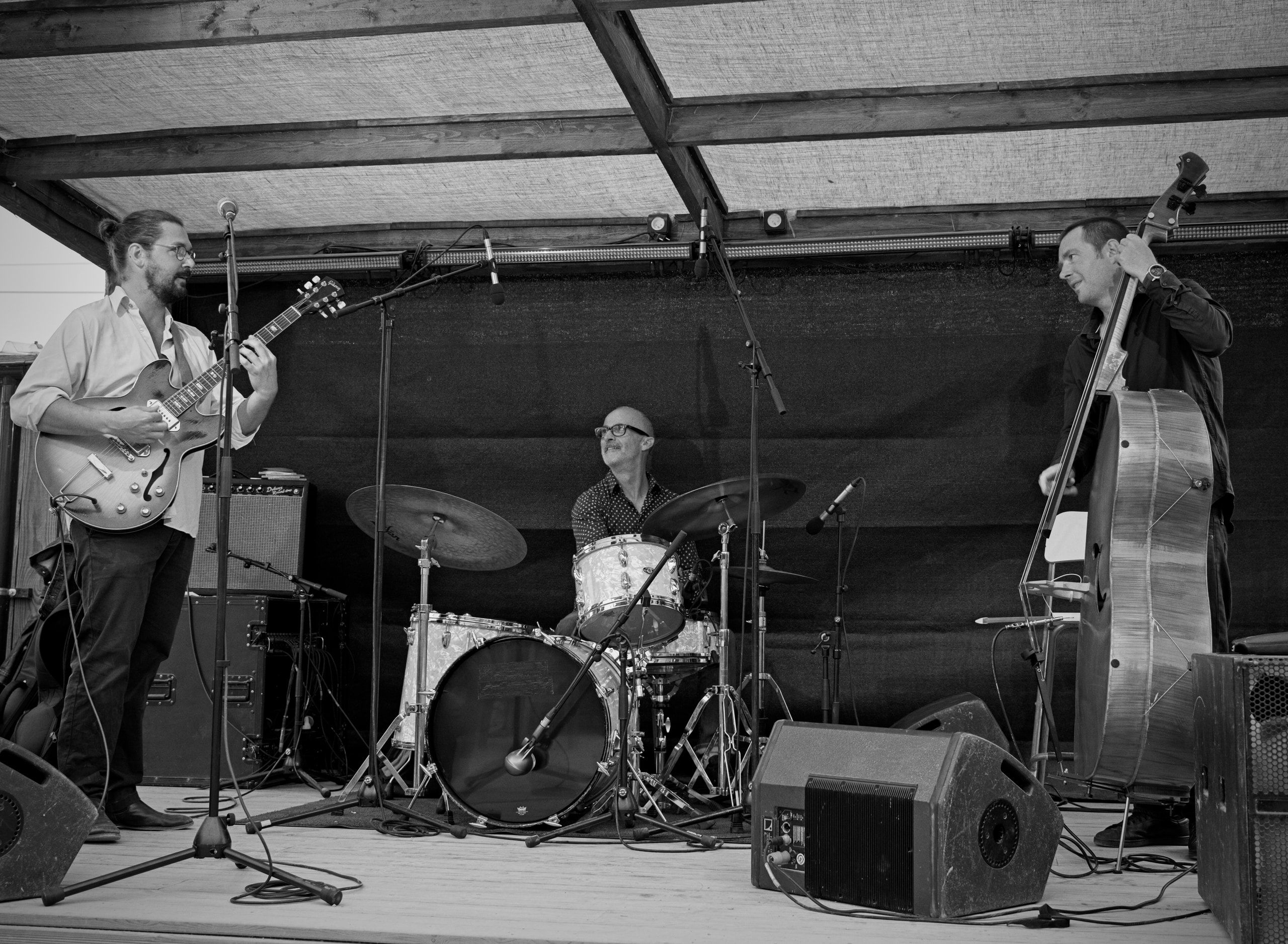 Le trio sur une scène en plein air, en noir et blanc : guitare, batterie et contrebasse.